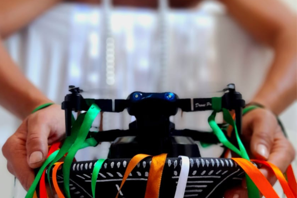Na cena central, uma mulher em transe carrega fogo e um drone como oferendas, enquanto outro drone com fitas brancas sobrevoa sua cabeça como guia espiritual